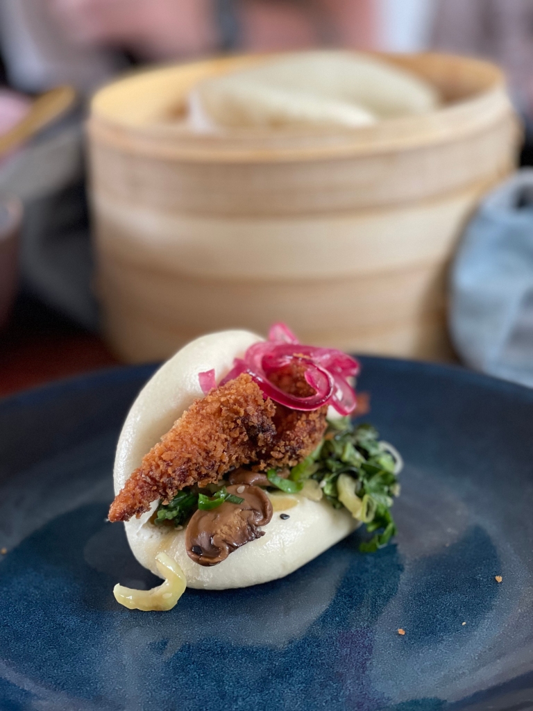 A filled bao bun sits on a deep blue plate. The pickled red onions in the bun contrast beautifully with the blue plate. In the background, a bamboo steamer fills the view.